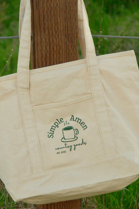close up of Beige canvas tote bag with a coffee cup and the text :"simple amen country goods est. 2025" screen printed on it on a wooden post against a grassy background