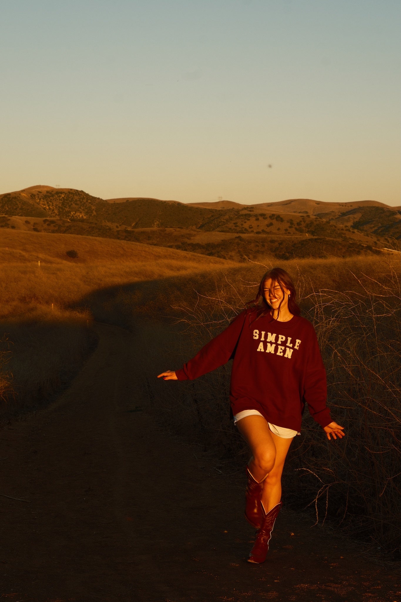 Person wearing a 'Simple Amen' sweatshirt with tan gingham patchwork text on the front saying "Simple Amen" in a desert landscape with warm lighting