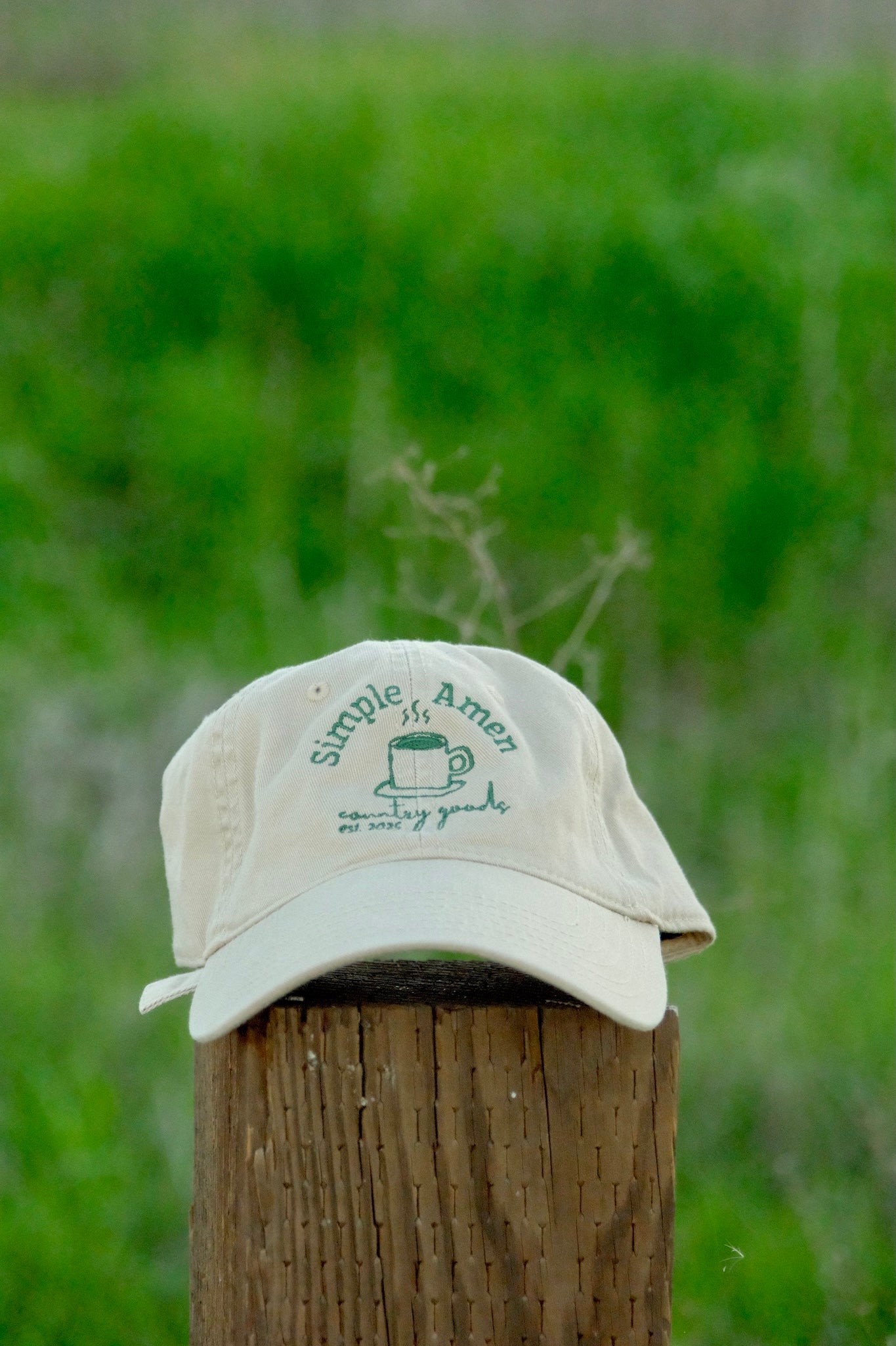 Beige cap with an embroidered green coffee cup, that says "simple amen country goods est. 2025 surrounding the cup" on a wooden post with green grass in the background