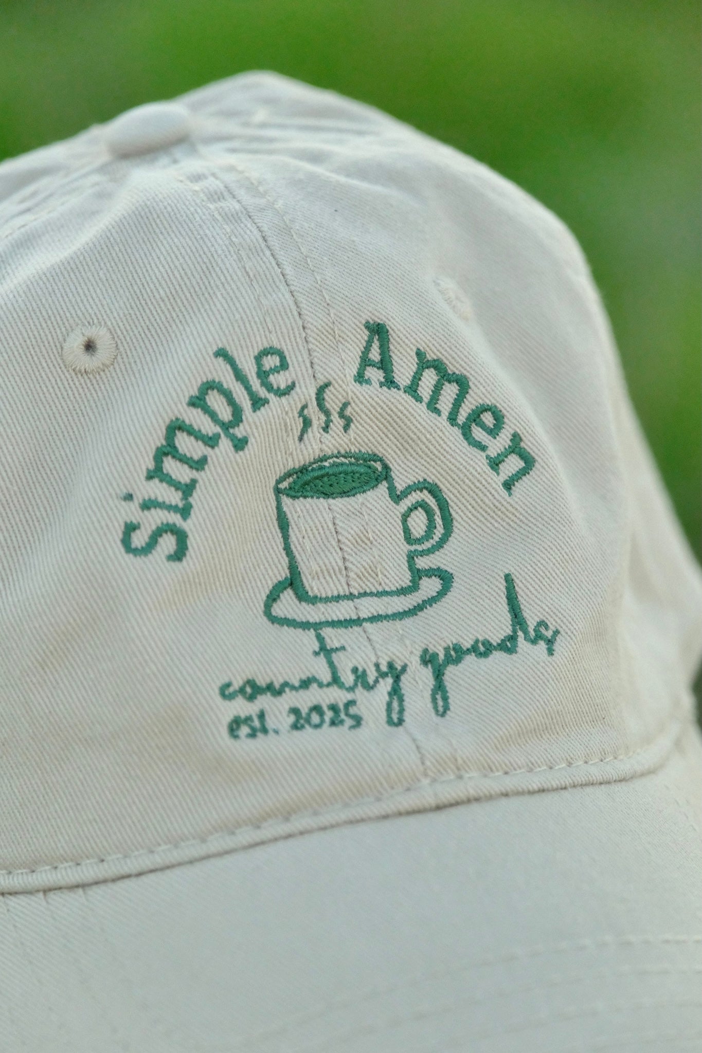 close up of Beige cap with an embroidered green coffee cup, that says "simple amen country goods est. 2025 surrounding the cup" on a wooden post with green grass in the background
