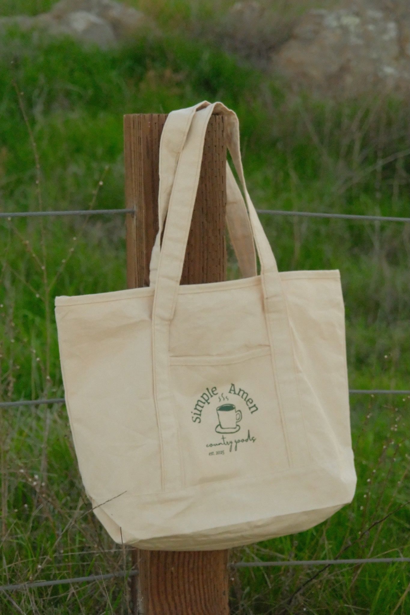 Beige canvas tote bag with a coffee cup and the text :"simple amen country goods est. 2025" screen printed on it on a wooden post against a grassy background