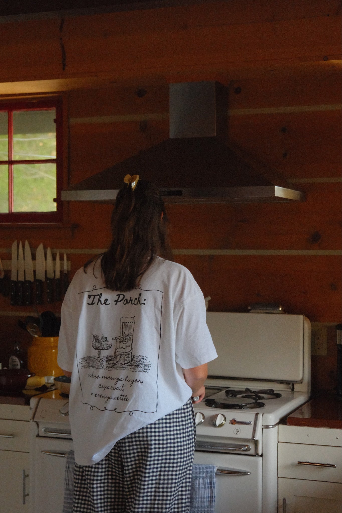 Woman wearing a white t-shirt with a black rocking chair and table. The tshirt features the words "The porch" on the top and "where mornings linger, cups await and evenings settle" on the bottom. This design is on the back. 
