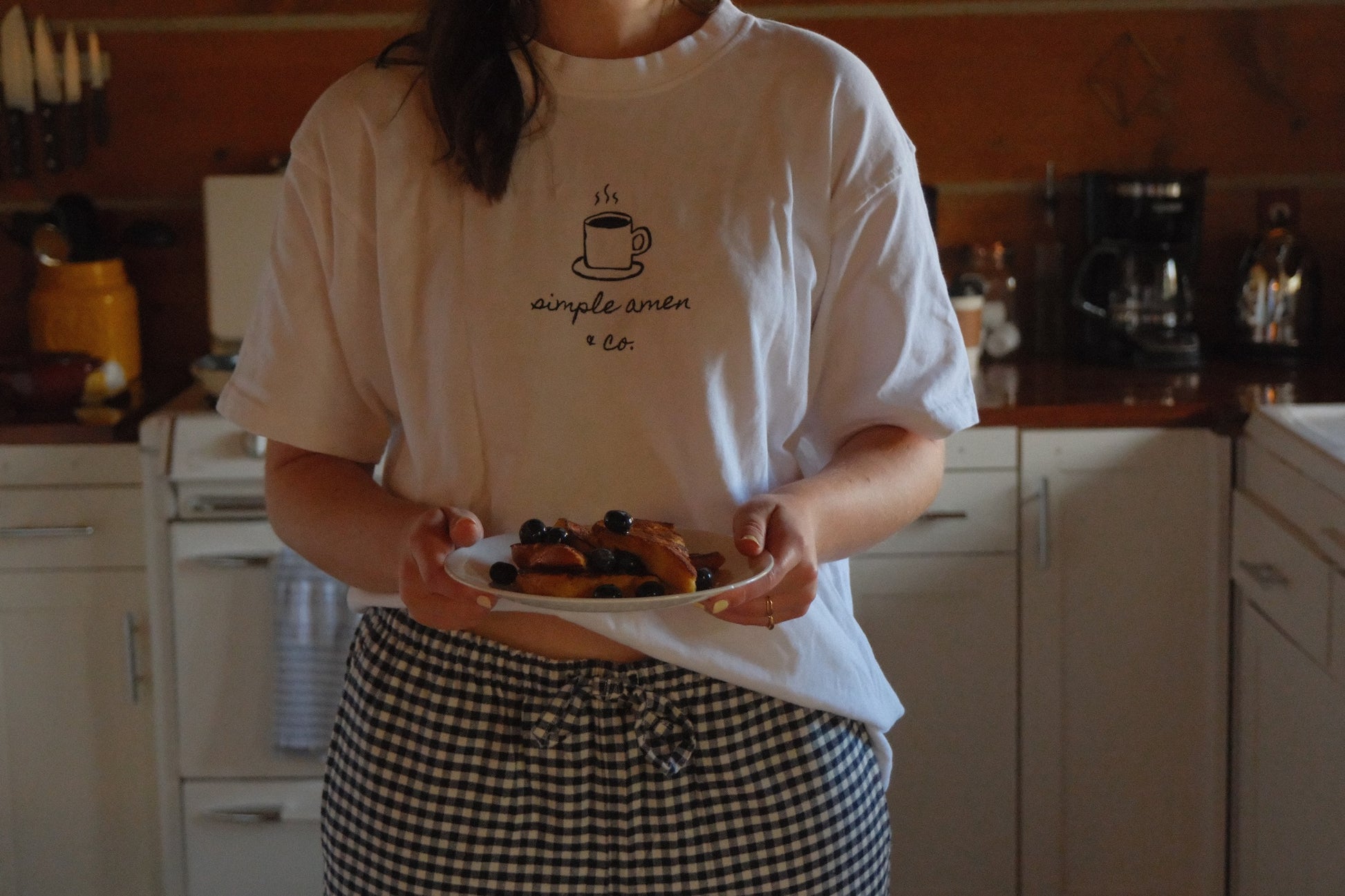 Person holding a plate of French toast with blueberries, wearing a white t-shirt with a coffee cup design and text saying "simple amen & co"