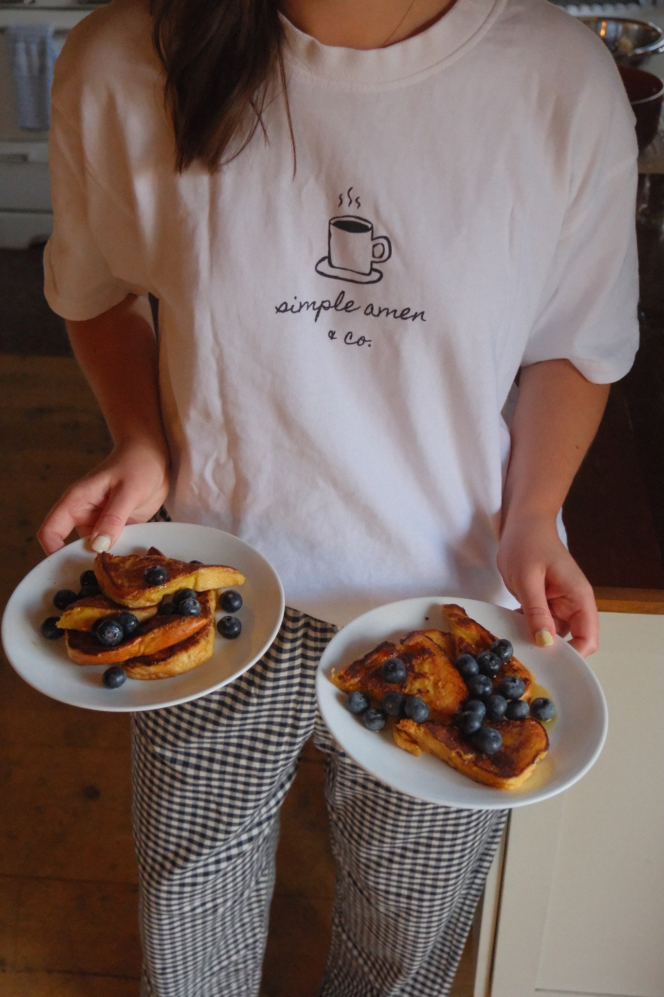 Person holding two plates of French toast with blueberries, wearing a white t-shirt with a coffee cup design and text saying "simple amen & co"