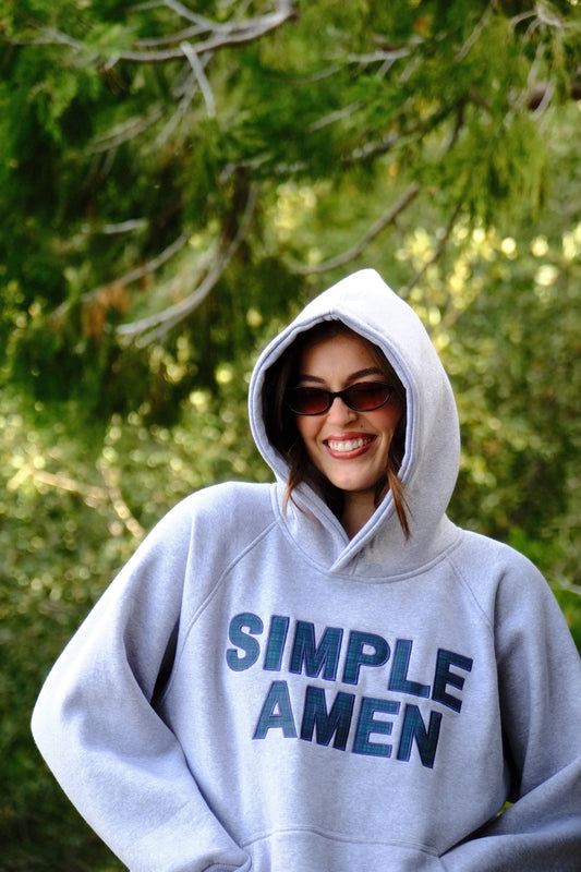 Person wearing a heather grey hoodie with 'Simple Amen' in green and blue plaid text on the front outdoors