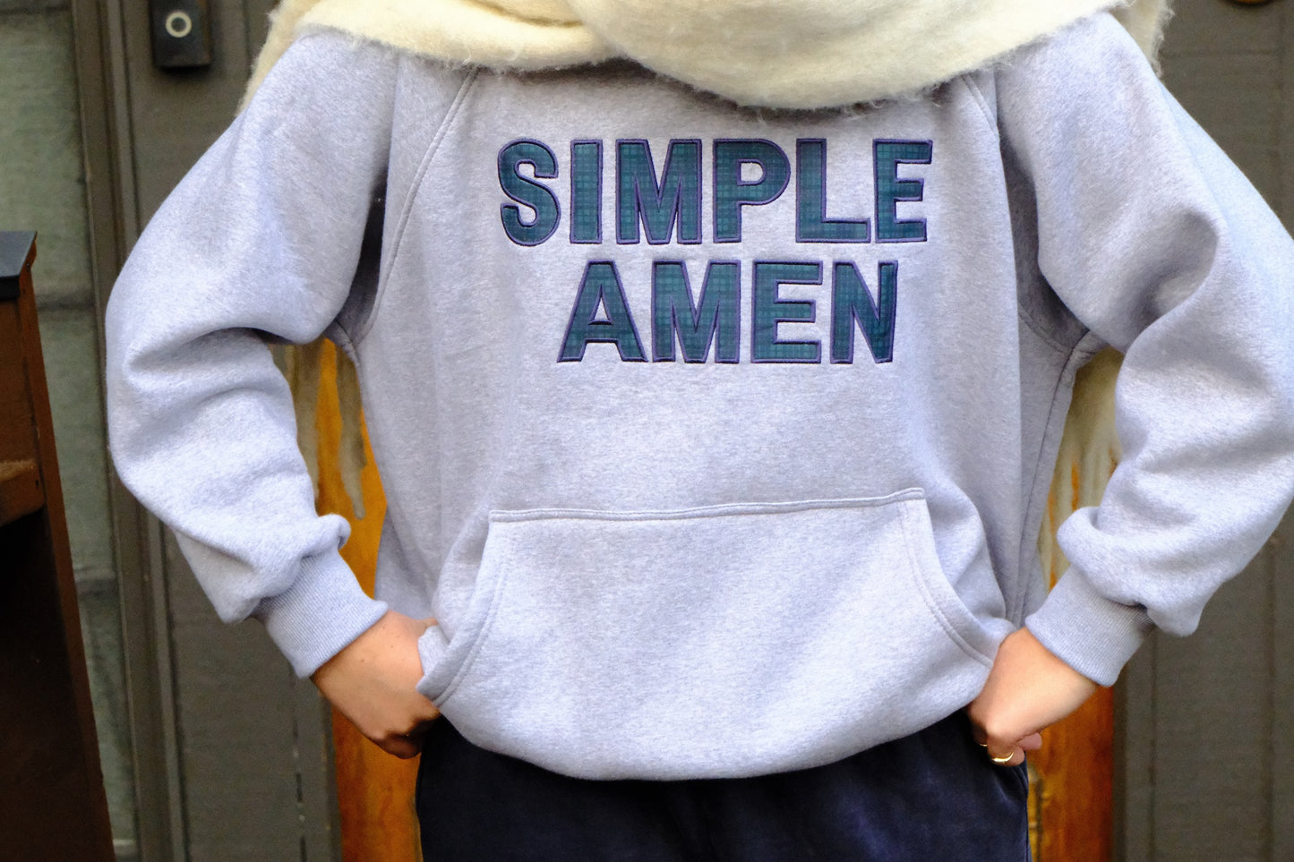 Person wearing a gray hoodie with 'SIMPLE AMEN' in blue and green plaid text on the front, standing in front of a door way
