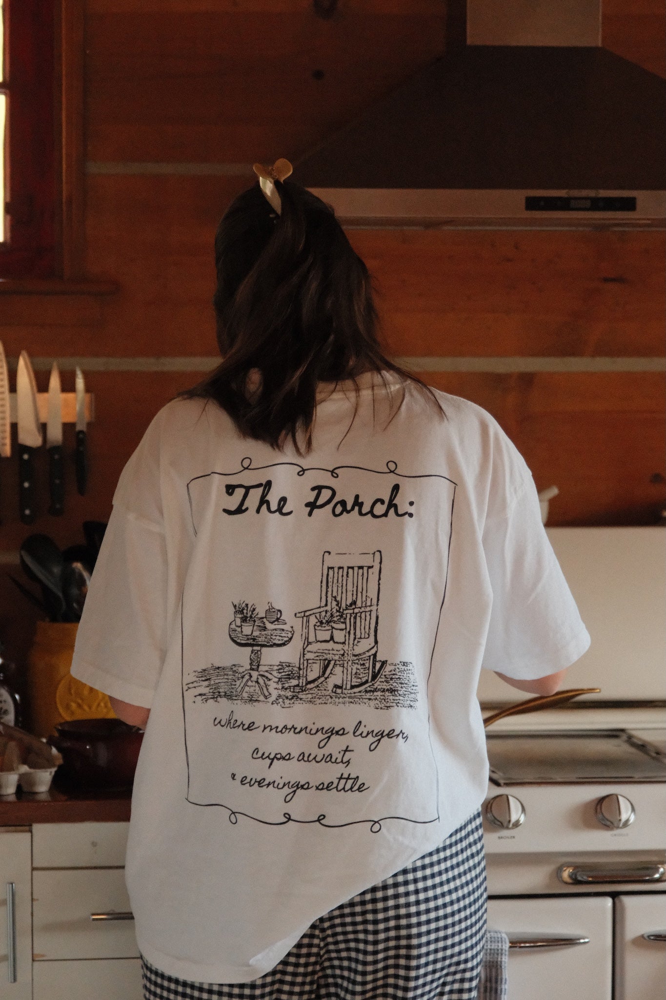 Woman wearing a white t-shirt with a black rocking chair and table. The tshirt features the words "The porch" on the top and "where mornings linger, cups await and evenings settle" on the bottom. This design is on the back. 